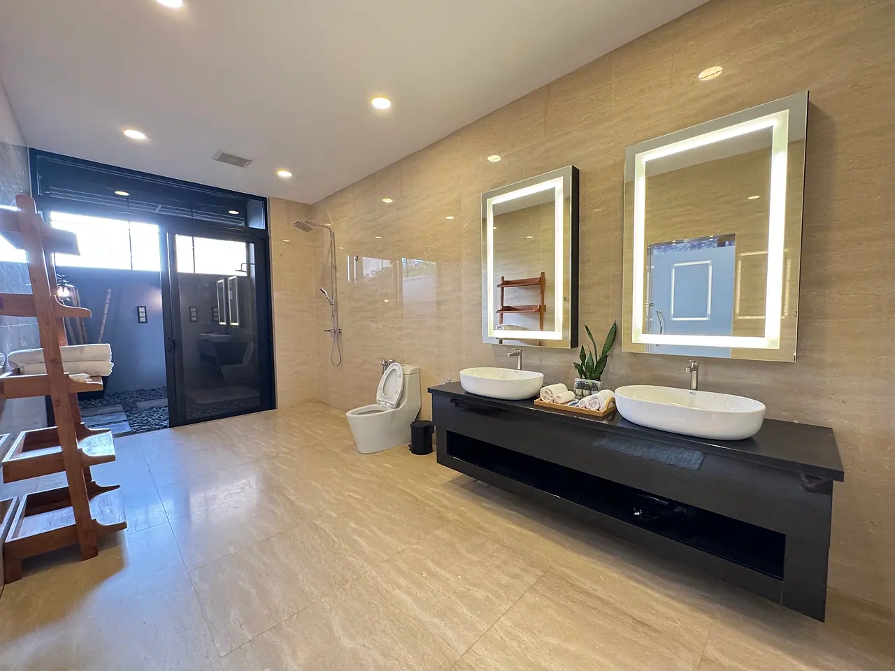 A deluxe bathroom: marble-look tiles, two basins under lit mirrors, and a glass door to the outdoor shower