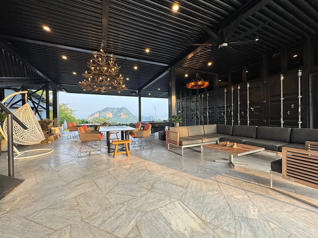 The open-air pavilion under its black steel roof: an antler chandelier, a long sofa and dining tables, with a limestone hill framed in the opening