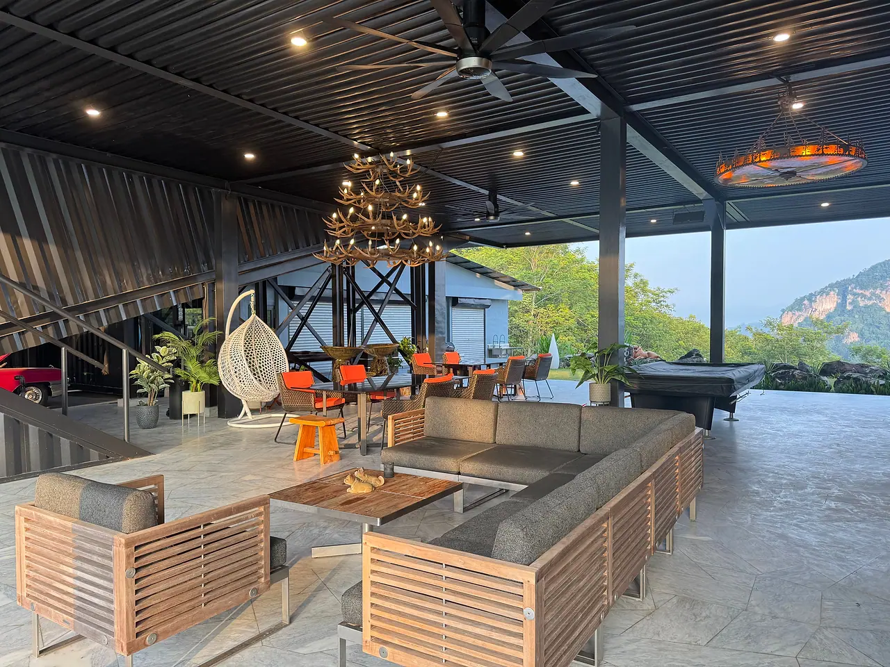 Teak sofas around a low table in the open-air pavilion, with a hanging egg chair, the pool table and a limestone hill beyond